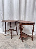 Two antique wooden hallway accent tables on concrete floor against white curtain. One kidney-shaped with turned legs, the other with carved slats and lower shelf.