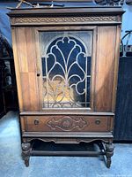Front view of the antique vintage wooden cabinet showing glass door with ornate wrought iron scrollwork and drawer with carved wood detail.