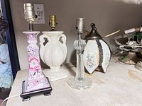 Four assorted vintage lamp bases on a table, including pink floral porcelain lamp, white porcelain vase-style lamp, clear ribbed glass lamp, and oval floral glass lamp with metal top.