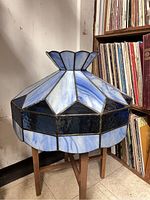 Blue stained glass lamp shade shown on a wooden stand, highlighting the geometric faceted panels and flower petal top design.