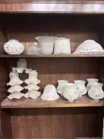 Lot of various glass lamp shades arranged on two shelves in a wooden cabinet. Includes ruffled frosted white shades, clear patterned, and floral decorated pieces.