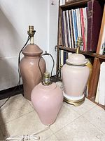 Three vintage pink tone ceramic table lamps of different sizes and shapes shown together on the floor with bookshelves in background.