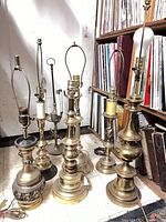Photo showing seven vintage brass lamps grouped on floor next to bookshelf. Lamps vary in design and size, all without shades, all untested.