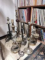 Photo showing a collection of eight vintage lamps arranged on the floor and in front of a bookshelf with vinyl records in the background. Visible details include brass, wood, glass components, and electrical cords.
