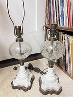 Pair of two vintage-style table lamps with white milk glass bases, metal footed bottoms, brass fittings, clear pressed glass globes with different patterns, cords and plugs visible.