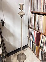 Full image of a tall rustic indoor floor lamp placed near a white door and a bookshelf filled with vinyl records. Lamp shows weathered finish and classical design elements.