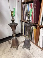 Photo of two antique metal table lamps side by side on floor against a bookshelf, showing overall shape and detailing of bases and lamp harps.