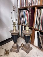 Pair of antique lamps positioned side by side showing entire lamp including base, midsection, and harp frame, with visible electrical cords on the floor.