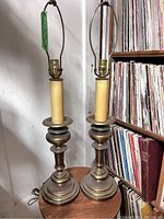 Pair of brass lamps on circular wooden table near shelving with records and books, showing full height and base design.