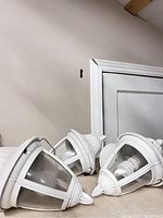 Three white outdoor wall sconces positioned on a surface, showing pointed tops and glass panels with bulbs inside.