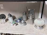 Photo showing a variety of vintage chrome wall sconces and light fixture parts arranged on a shelf with visible glass shades, chrome bases, mounting hardware, and exposed wires.