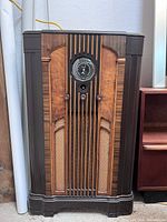 Front view of vintage floor radio cabinet showing wood and fabric design, dial housing, and control knobs.