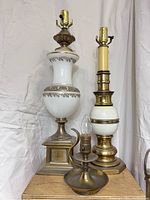 Photo showing three vintage table lamps together on a wooden surface: one large porcelain and brass with floral design, one tall round brass and white base lamp, and one smaller brass lamp with exposed bulb.