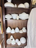 Photo showing multiple shelves with various white glass lamp shades in different shapes and sizes arranged on a wooden shelving unit.