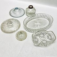 Six antique glassware pieces arranged on white surface including covered dishes, serving trays, and an ink bottle.