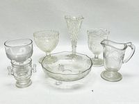 Six clear antique glassware items arranged in front of a plain white background, including goblets, a vase, a bowl, and a pitcher, showing various etched and pressed patterns on the glass.