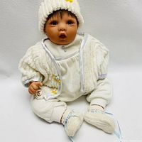 Front view of lifelike baby doll seated against white background wearing white knit hat with yellow flower, white textured jacket with satin trim, white onesie, pants, and knit booties.