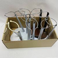 Photo of eight doll stands in a cardboard box showing various metal ring designs and bases. Mostly silver metal stands with one wooden base.
