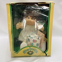 Front view of Cabbage Patch Kids doll inside yellow and green box with torn front window plastic and box damage.
