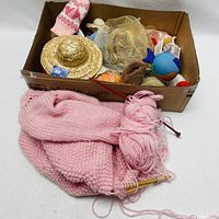 View of box containing assorted folk art and craft items, and pink knitted scarf with yarn and needles in front of box.
