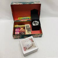 Jewelry box open showing floral interior with magnetic jewelry clasp pack, decorative lighters, large faceted faux crystal in black box, and a blue pen box.