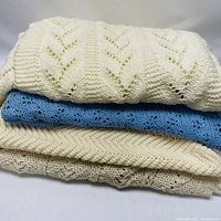 Stack of three knit throws and bedspreads showing cream and blue colors and knit patterns.