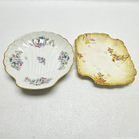 Two Limoges plates, shell-shaped and square, with floral patterns and gold trim on a white surface.