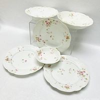 Top view showing six Limoges porcelain items: three scalloped rim bowls on risers, small scalloped bowl, large serving plate, and dinner plate all decorated with pink floral patterns on white porcelain.