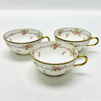 Three Limoges porcelain teacups decorated with pink roses, purple ribbons, green leaves, and trimmed in gold on rims and handles.