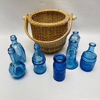 Front view showing six blue miniature glass bottles with varying shapes and embossed details next to small woven basket with wooden handles.