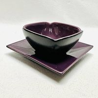 Purple ceramic bowl with scalloped edges on matching square plate against a white backdrop.