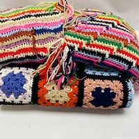 Two folded colorful crochet throws stacked, showing varied multicolored granny square motifs and stripe patterns with fringed yarn tassels on edges.