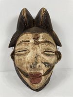Front view of the African Okuyi helmet mask showing facial details including closed eyes, painted white face with black markings, and raised hairstyle details.