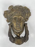Front view of brass door knocker featuring a stylized female face with decorative elements around the head and a ring knocker below.