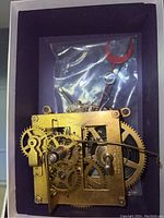 Brass clock mechanism with visible gears and springs, some markings of E Ingraham & Co. Packaging shows red pendulum part behind mechanism.
