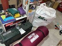 Overview of camping items showing stove in green case, boxed propane heater, tent box, sleeping bag in storage sack and smaller green and purple bags