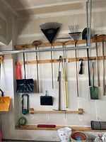 Wide view of garden tools hanging on a wooden rack in the garage, showing various rakes, shovels, hoes, and snow shovels.