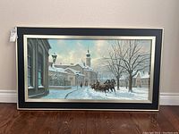Framed winter scene painting by Georges Csapo showing a snowy village street with horses and passengers.