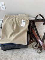 Folded beige khaki pants stacked on other pants next to a cluster of several leather belts in brown and black.