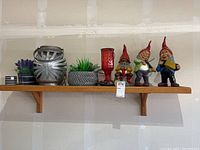 Whole set of items on a wooden shelf including 3 garden gnomes, 2 artificial plants, 1 lantern, 1 candle holder, and 1 small planter.