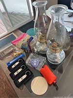 Two glass oil lamps, Egg Genie in packaging, three empty glass cases, wallet, handheld fan, salt shaker, multiple keychains including laundry bag shaped and pink leather keychain, round wooden coaster/base on table.