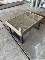 Full view of rectangular outdoor coffee table showing glass top, wicker sides, and black metal legs on concrete floor