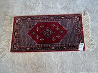 Full view of small rectangular red Tunisian silk rug with fringes and geometric central design.
