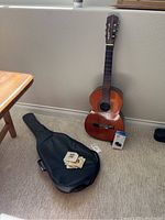 Full view of Suzuki acoustic guitar leaning against wall with black soft guitar case lying on floor and extra accessories around it.