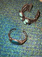 Two silver-tone cuff bracelets with turquoise stones displayed on textured blue-green surface.