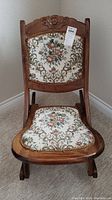 Front view of vintage small folding rocker chair showing floral upholstery and carved wood detail on backrest