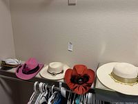 Five women’s hats displayed on a shelf: pink cowboy hat, white cowboy hat, red cowboy hat with feather, and two large brim hats, plus two hat boxes.