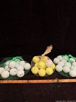 Full lot showing three mesh bags: two bags contain white golf balls, one bag contains yellow golf balls. A wooden ruler is placed in front for scale.