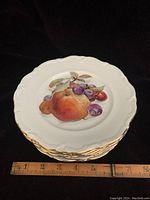 Stack of 6 dessert plates with fruit design and gold rim shown with ruler for scale.