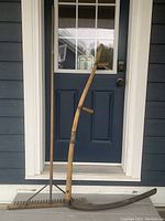 Full view of antique wooden grim reaper scythe and primitive wood hay rake placed against a door for scale.